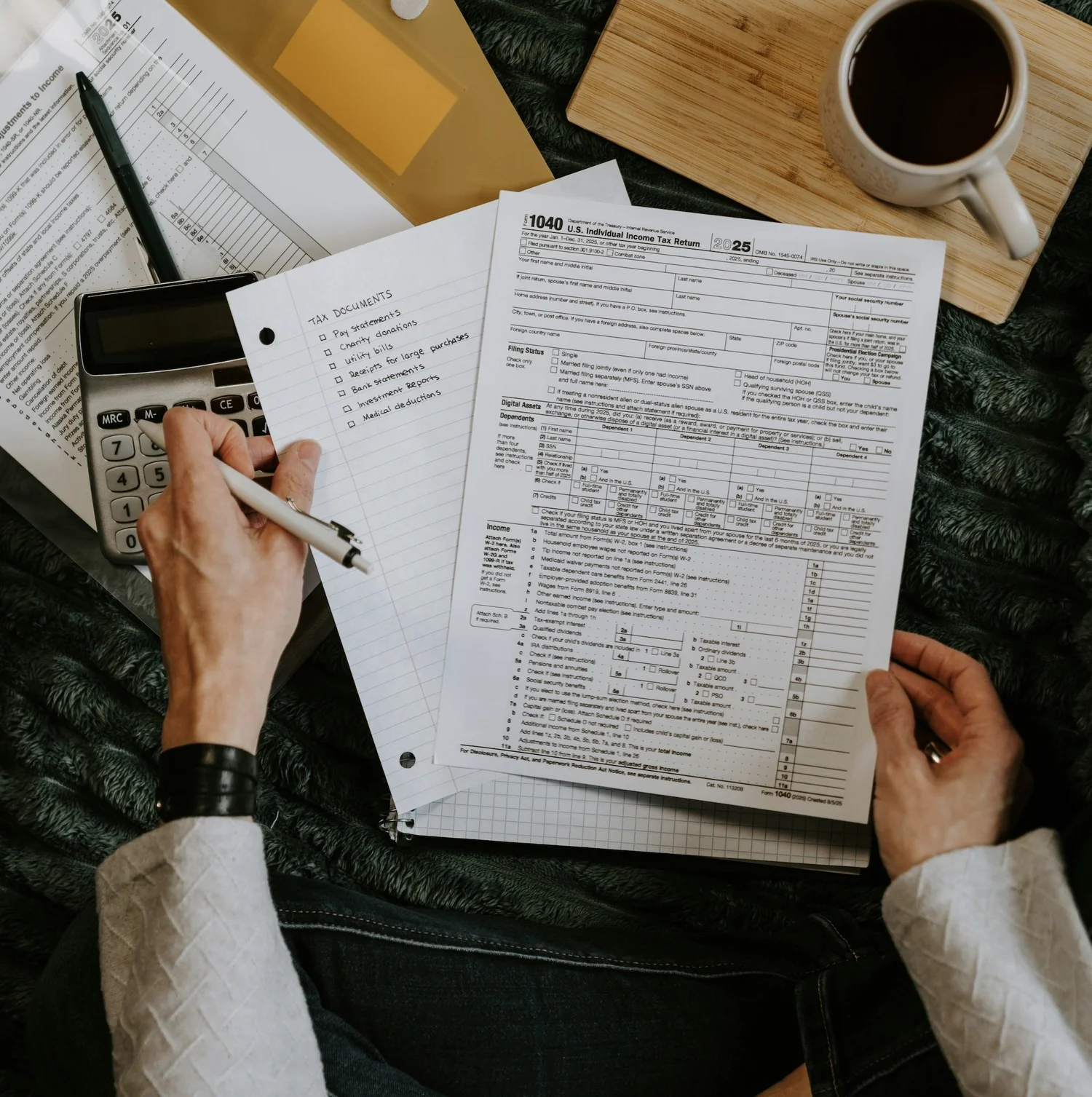 Hand filling out a 1040 tax form beside a calculator and coffee